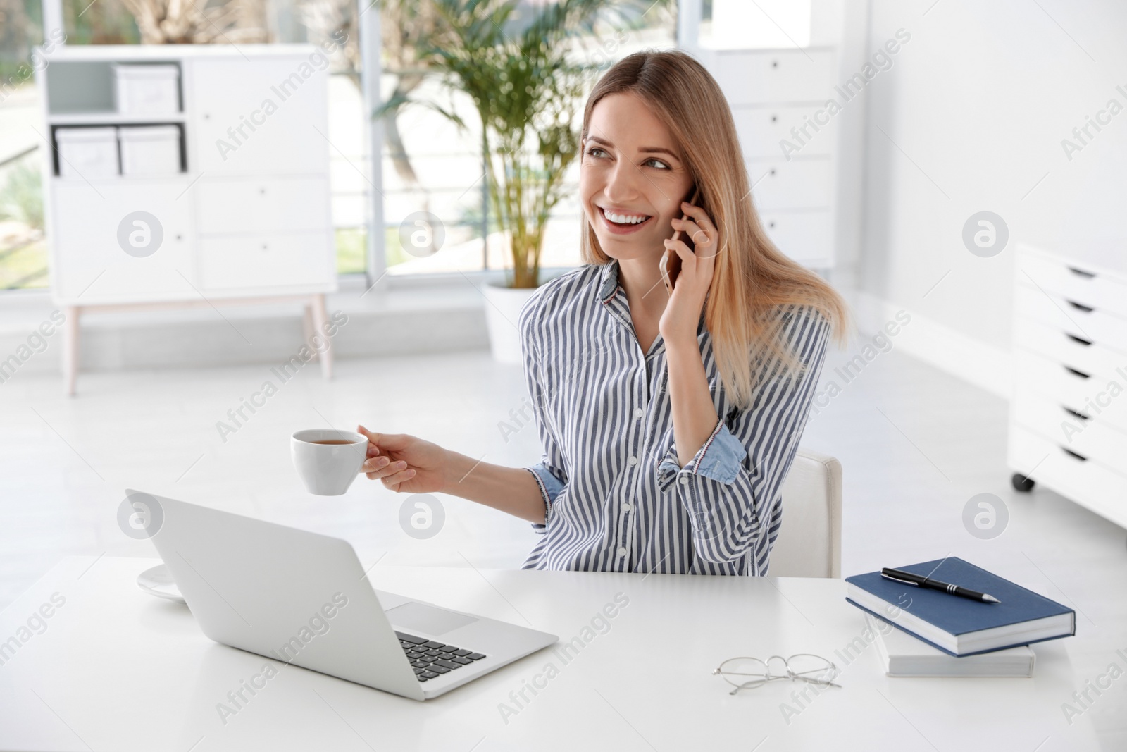 Photo of Young businesswoman talking on phone while using laptop at table in office