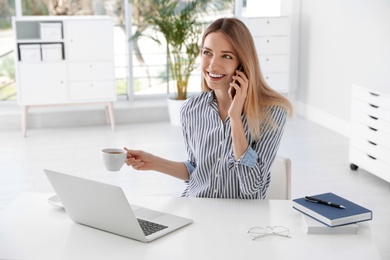 Young businesswoman talking on phone while using laptop at table in office