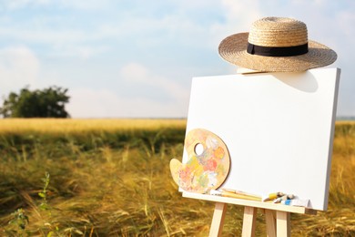 Wooden easel with blank canvas, painting equipment and hat in field. Space for text