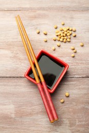 Photo of Soy sauce in bowl, soybeans and chopsticks on wooden table, flat lay