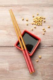 Soy sauce in bowl, soybeans and chopsticks on wooden table, flat lay