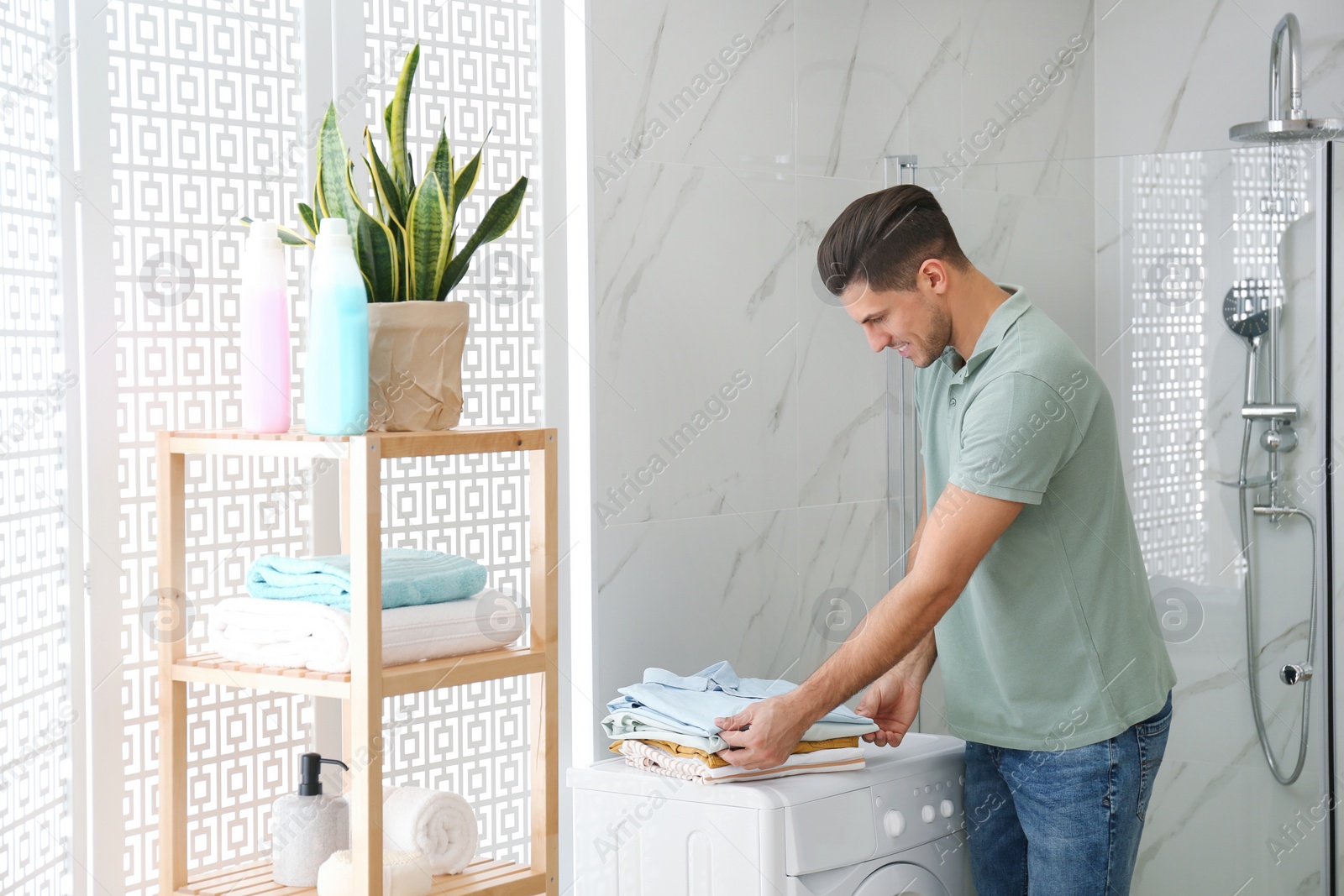 Photo of Happy man with clean clothes near washing machine in bathroom. Laundry day