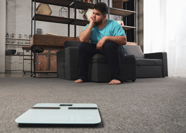 Photo of Depressed overweight man looking at scales in living room