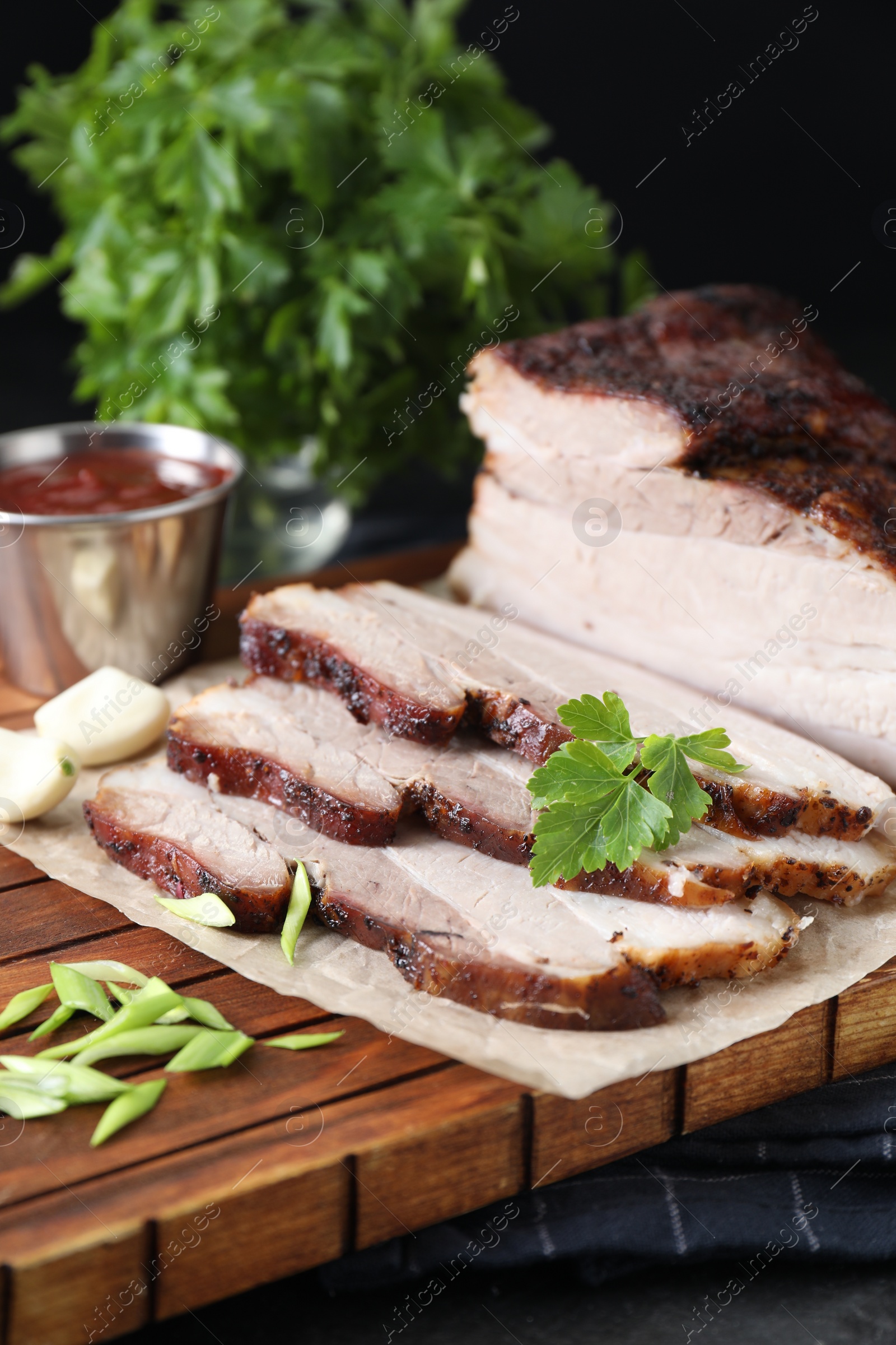 Photo of Pieces of baked pork belly served with sauce and parsley on black textured table, closeup