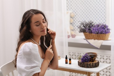 Photo of Beautiful young woman combing hair after applying essential oil at table indoors