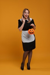 Photo of Emotional woman in scary maid costume with pumpkin bucket on orange background. Halloween celebration