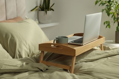 Photo of Wooden tray with modern laptop and cup of aromatic drink on bed indoors