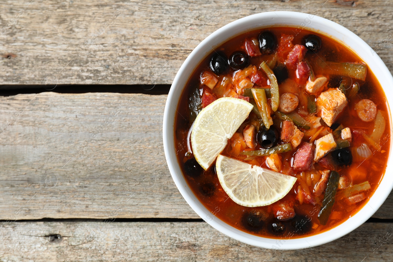 Photo of Meat solyanka soup with sausages, olives and vegetables in bowl on wooden table, top view. Space for text