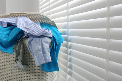 Plastic laundry basket overfilled with clothes near window blinds indoors, closeup. Space for text