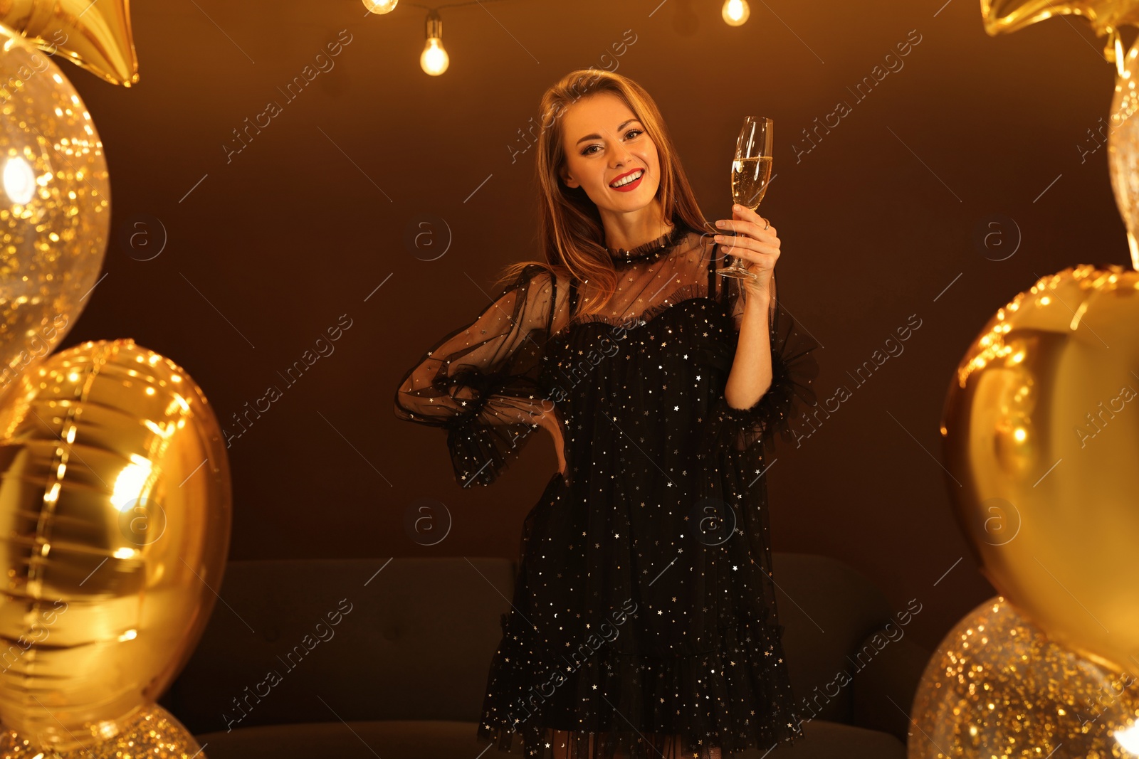 Photo of Beautiful woman with glass of sparkling wine near golden balloons indoors. New Year celebration