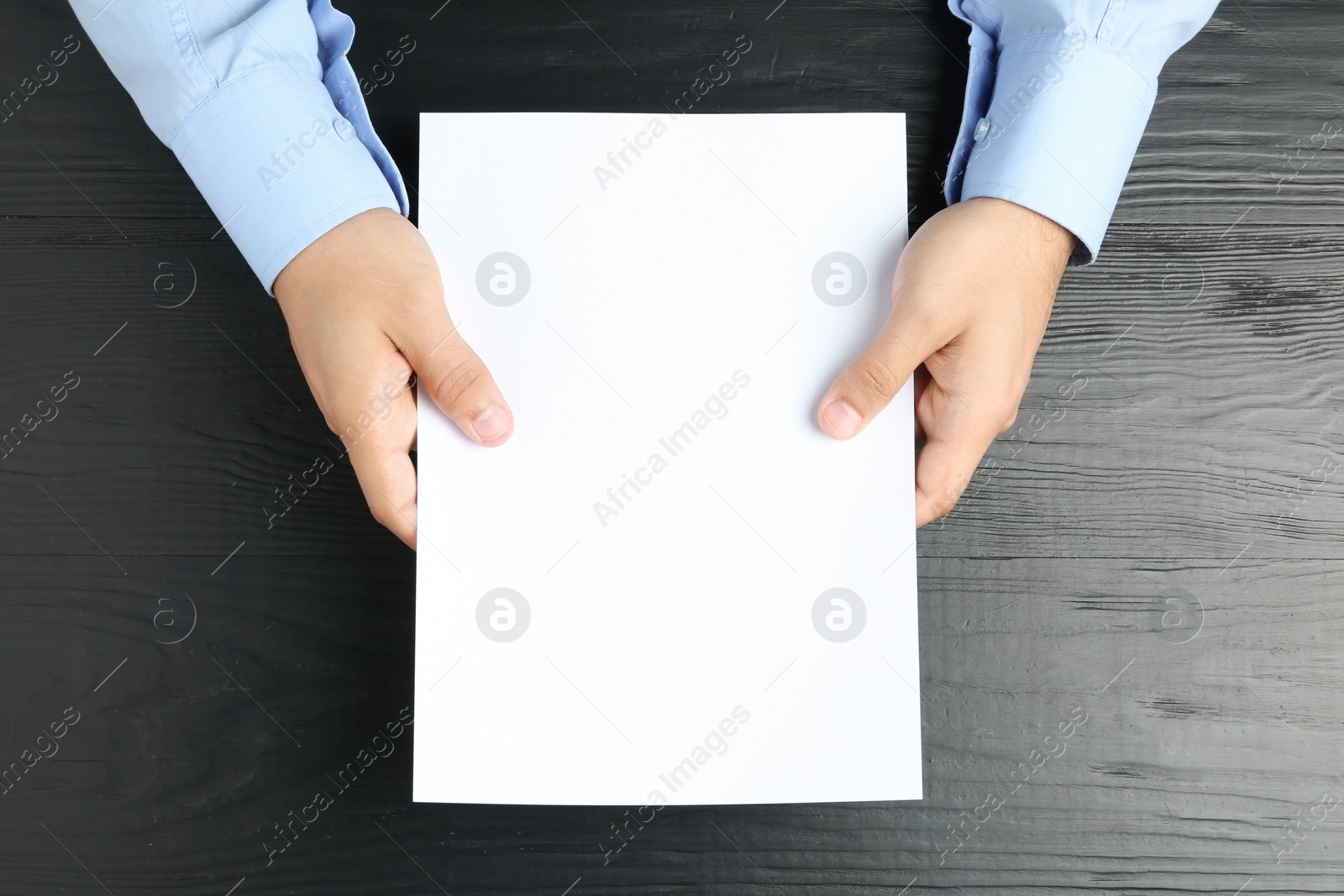 Photo of Man holding blank paper sheet for brochure at black wooden table, top view. Mock up