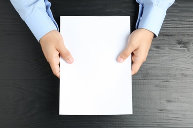 Man holding blank paper sheet for brochure at black wooden table, top view. Mock up