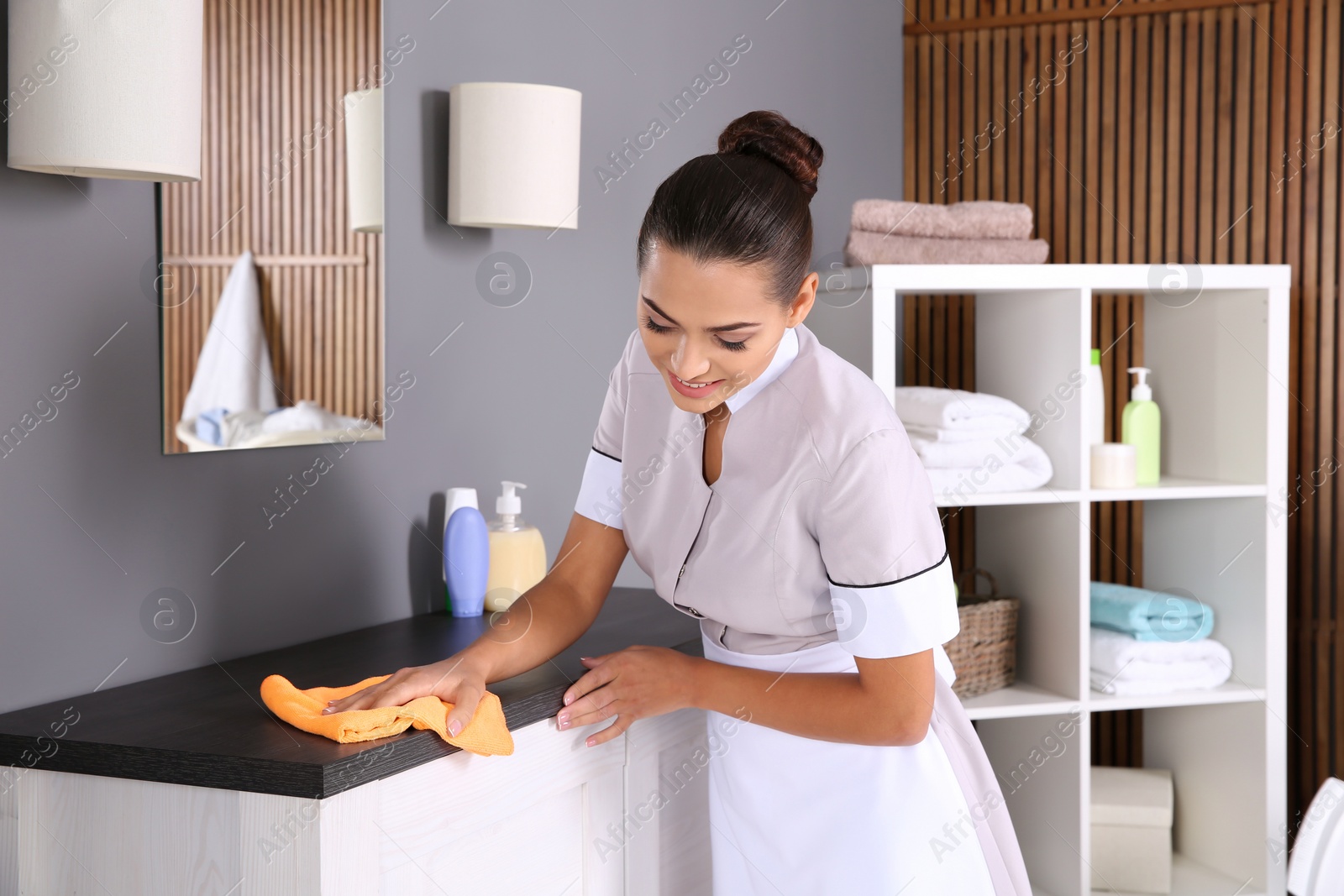 Photo of Young chambermaid wiping dust from furniture with rag in bathroom