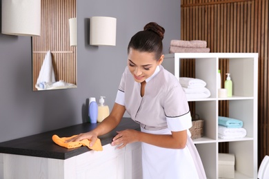 Photo of Young chambermaid wiping dust from furniture with rag in bathroom