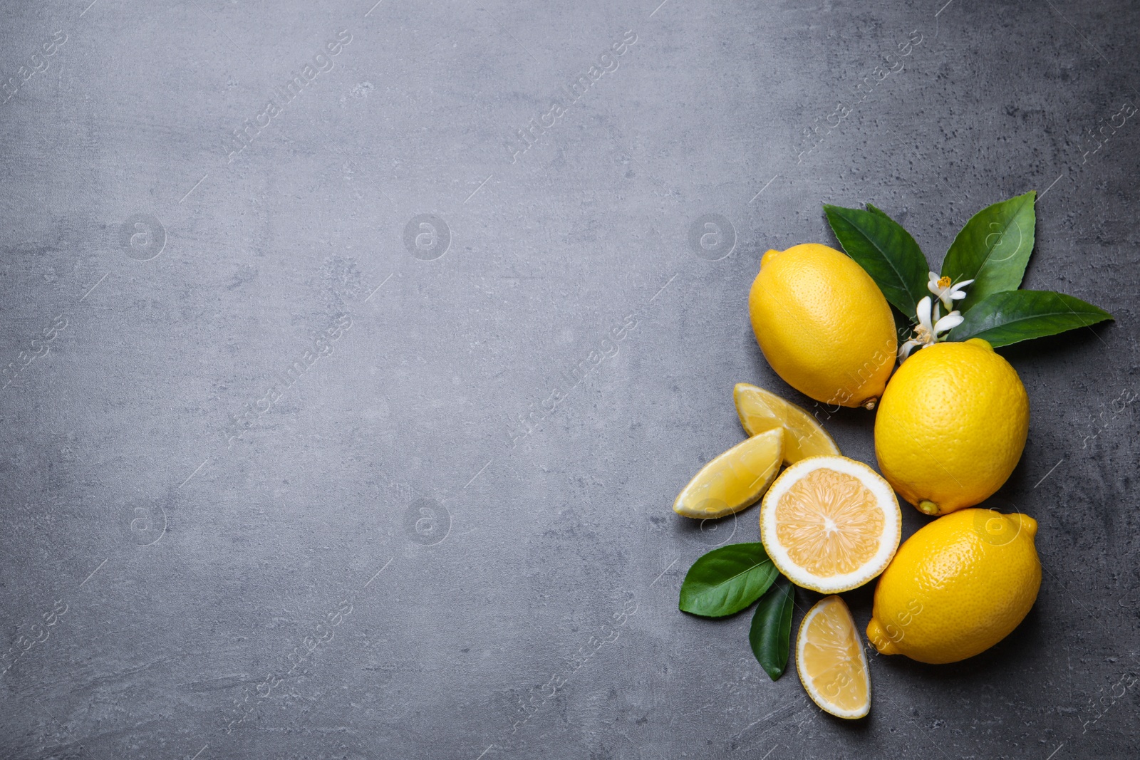 Photo of Many fresh ripe lemons with green leaves and flowers on grey table, flat lay. Space for text