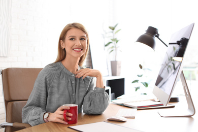 Young woman with cup of drink relaxing at table in office during break