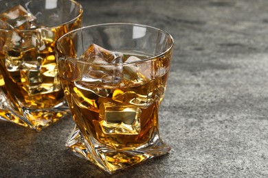 Whiskey and ice cubes in glasses on grey textured table, closeup. Space for text