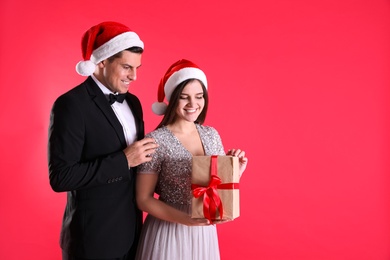 Beautiful happy couple in Santa hats holding Christmas gift on red background. Space for text