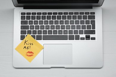 Photo of Sticky note with phrase Kiss Me and lipstick mark on laptop at white wooden table, top view