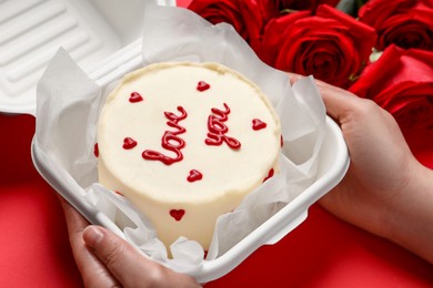 Photo of Woman holding takeaway box with bento cake at red table, closeup. St. Valentine's day surprise