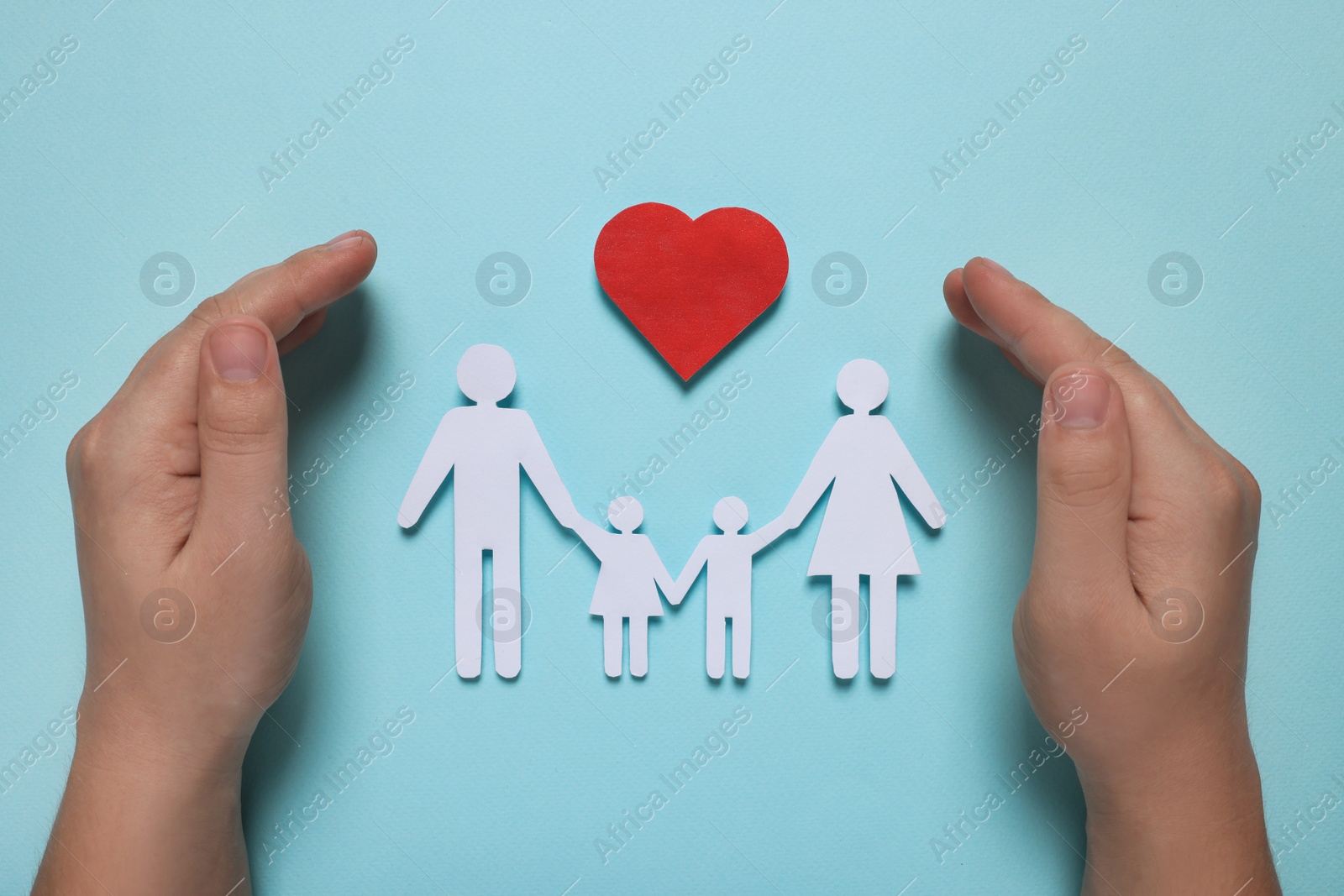 Photo of Man protecting paper family figures and red heart on light blue background, top view. Insurance concept