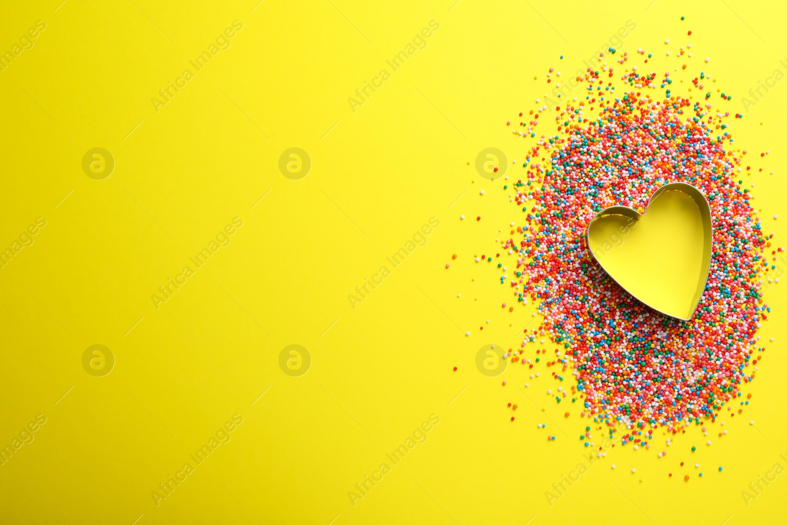 Photo of Heart shaped cookie cutter and sprinkles on yellow background, flat lay with space for text. Confectionery decor