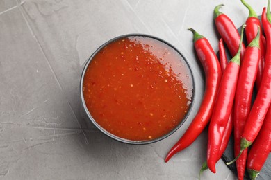 Photo of Spicy chili sauce in bowl and fresh peppers on grey table, flat lay