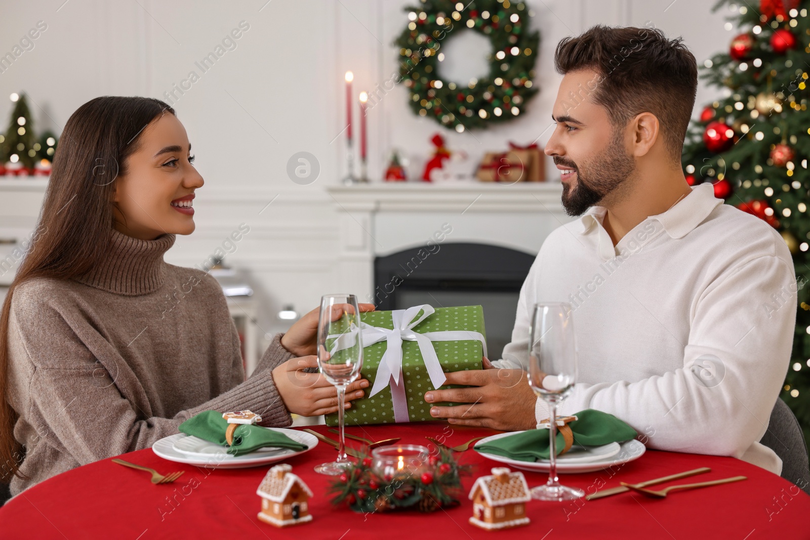 Photo of Happy young man presenting Christmas gift to his girlfriend at table indoors