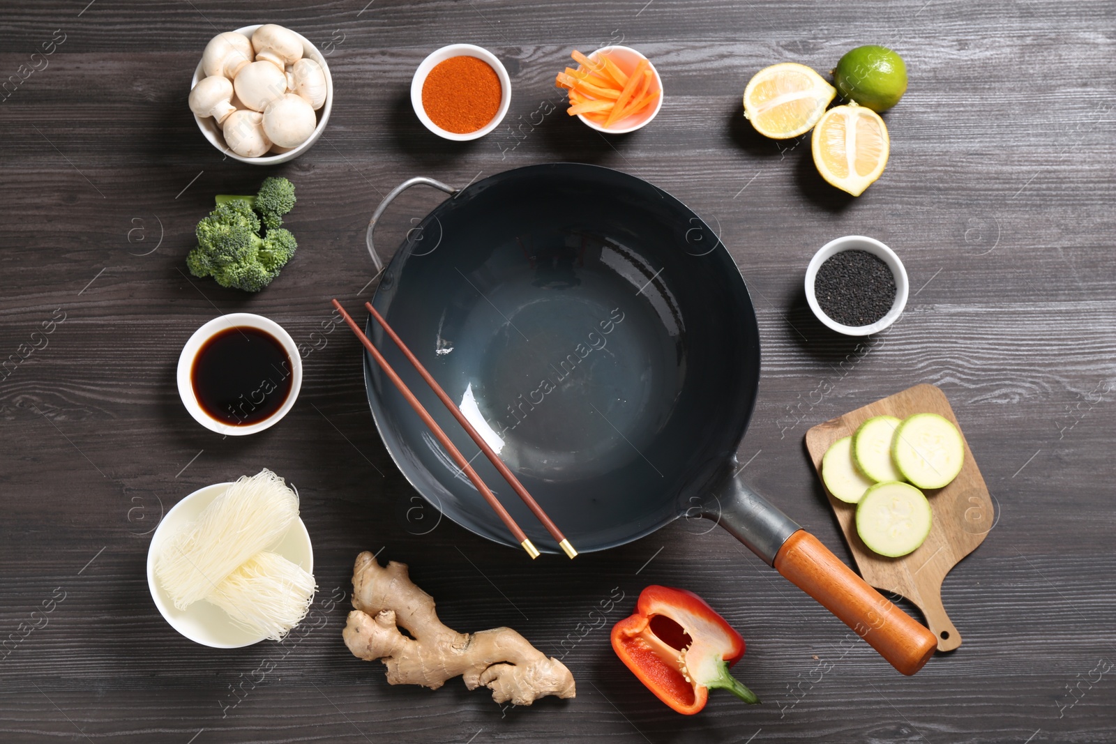 Photo of Empty iron wok and chopsticks surrounded by ingredients on dark grey wooden table, flat lay