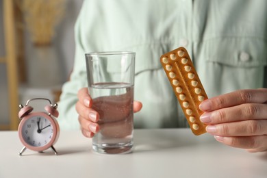 Photo of Woman holding blister of oral contraceptive pills and glass with water at white table, closeup