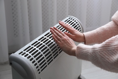 Photo of Woman warming hands near electric heater at home, closeup