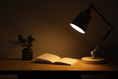 Photo of Stylish modern desk lamp, open book and plant on wooden table near wall in dark room