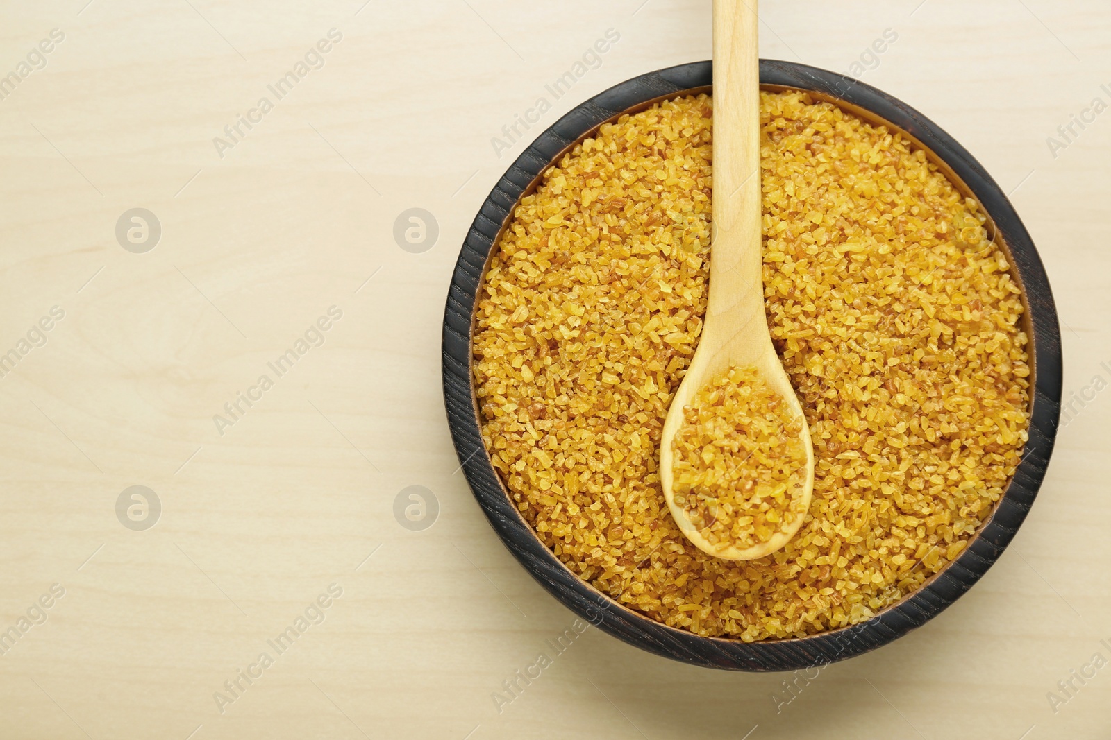 Photo of Bowl and spoon of uncooked bulgur on wooden table, top view