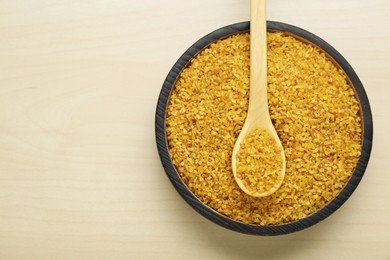 Bowl and spoon of uncooked bulgur on wooden table, top view