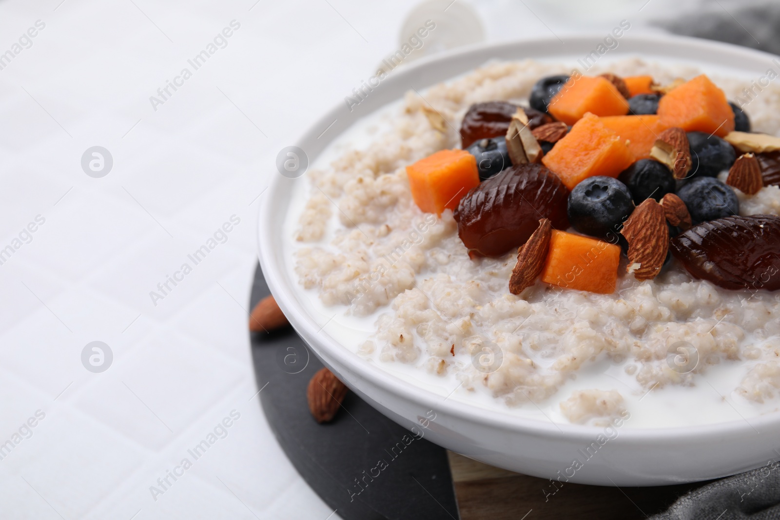 Photo of Delicious barley porridge with blueberries, pumpkin, dates and almonds in bowl on white tiled table, closeup. Space for text