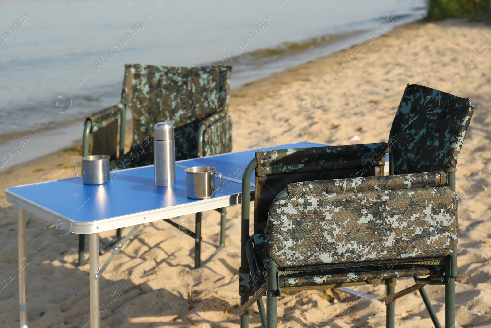 Photo of Camouflage fishing chairs and table with metal cups on sandy beach near river