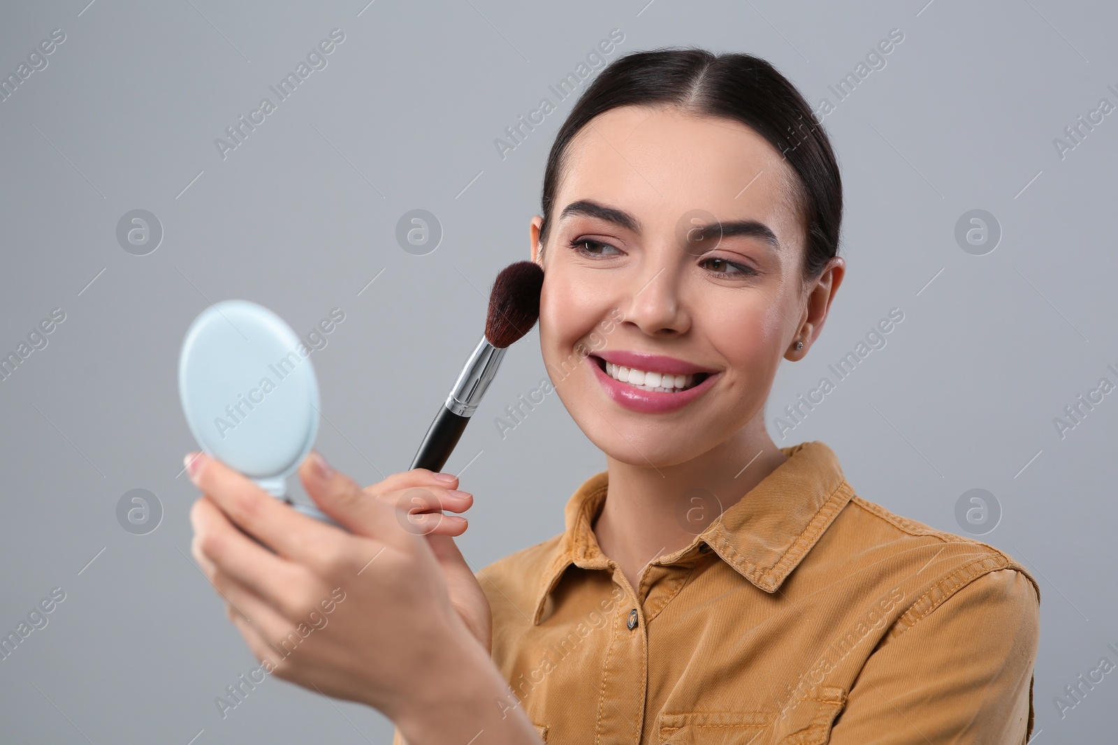 Photo of Happy woman with cosmetic pocket mirror applying makeup on light grey background