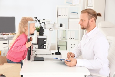 Photo of Children's doctor examining little girl with ophthalmic equipment in clinic