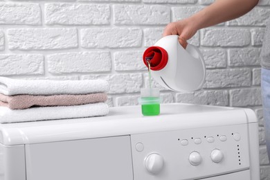 Photo of Woman pouring fabric softener from bottle into cap on washing machine near white brick wall, closeup