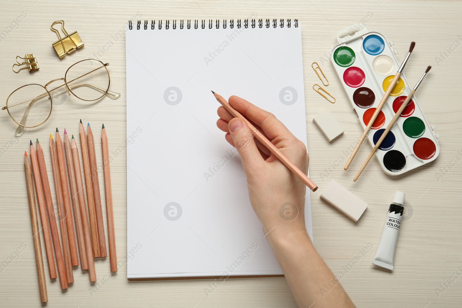 Photo of Woman with pencil and blank sketchbook at white wooden table, top view