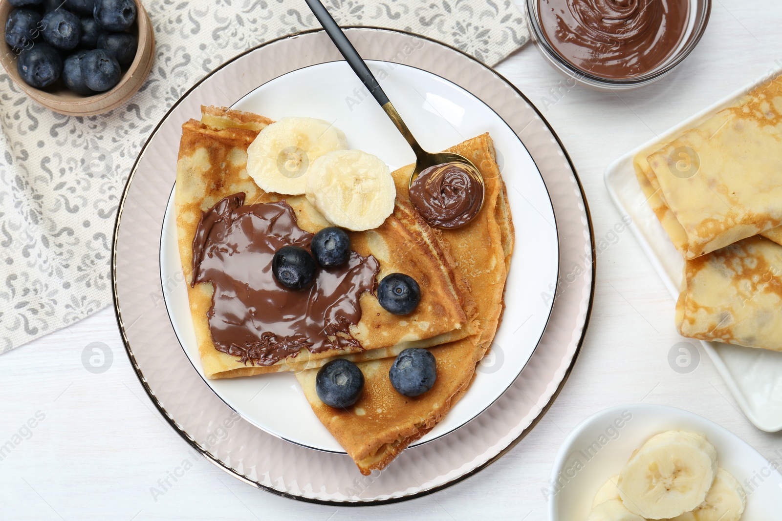 Photo of Tasty crepes with chocolate paste, blueberries and banana served on white wooden table, flat lay