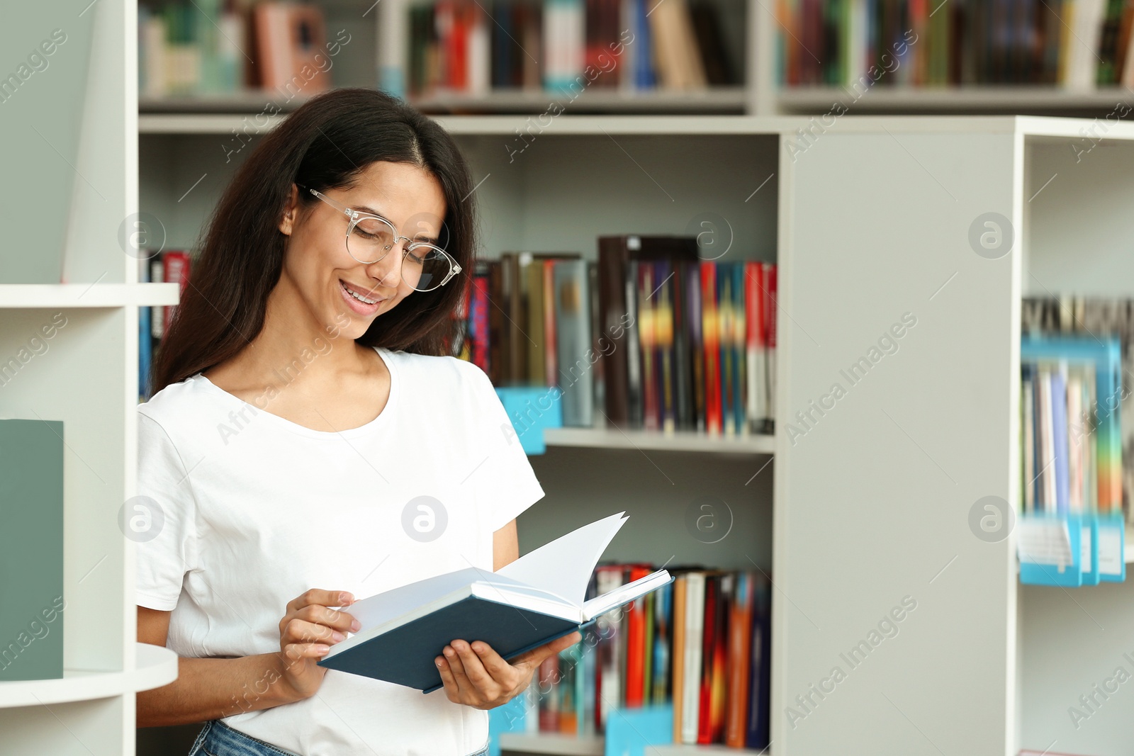 Photo of Young woman with book near shelving unit in library. Space for text