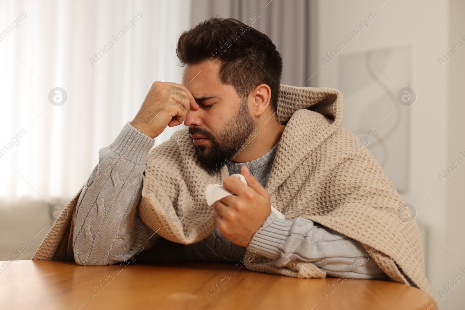 Photo of Sick man wrapped in blanket with tissue at wooden table indoors. Cold symptoms