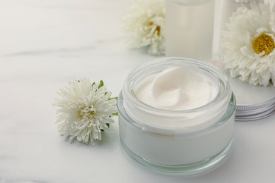 Photo of Glass jar of face cream and flowers on white marble table. Space for text