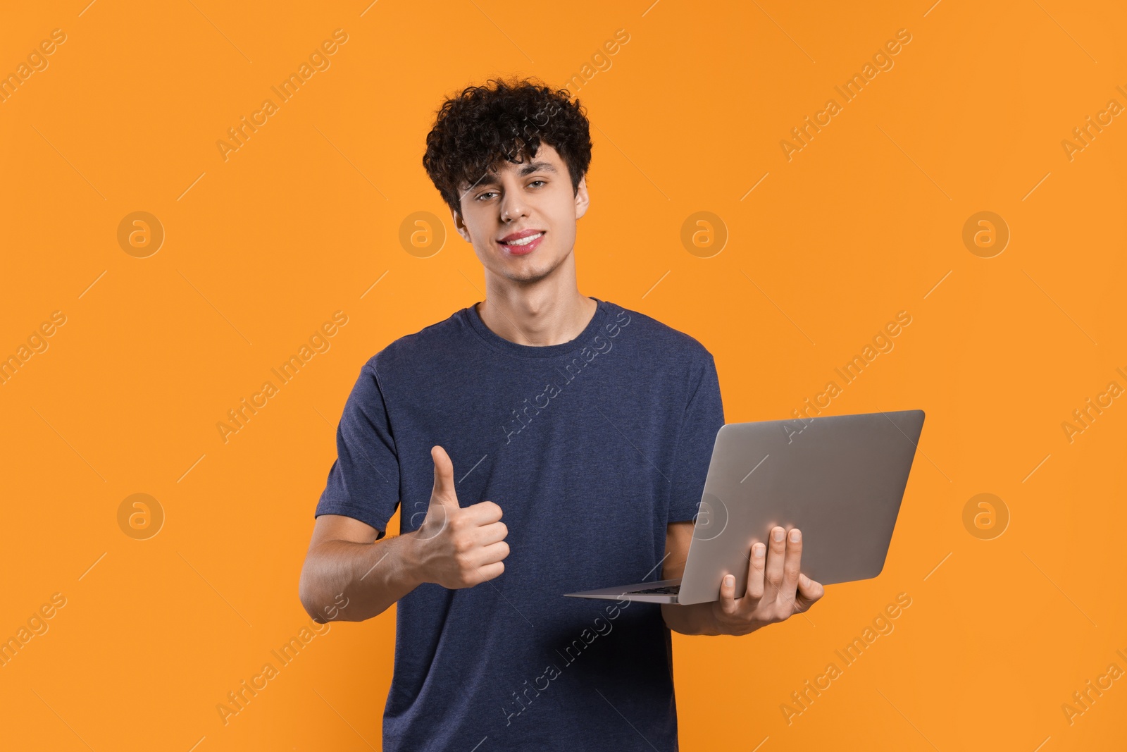 Photo of Handsome young man with laptop showing thumb up on orange background