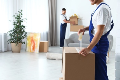 Photo of Moving service employee sealing cardboard box with adhesive tape in room, closeup