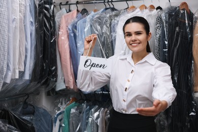 Photo of Dry-cleaning service. Happy worker holding Open sign near rack with clothes indoors, space for text