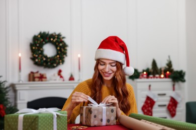 Photo of Beautiful young woman in Santa hat decorating Christmas present at table indoors