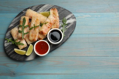 Photo of Tasty fried spring rolls, arugula, lime and sauces on light blue wooden table, top view. Space for text