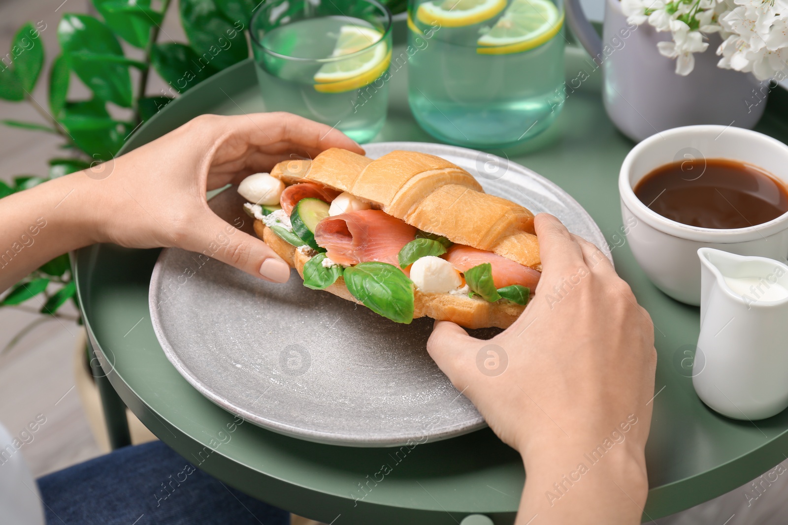 Photo of Woman holding tasty croissant sandwich over plate at table